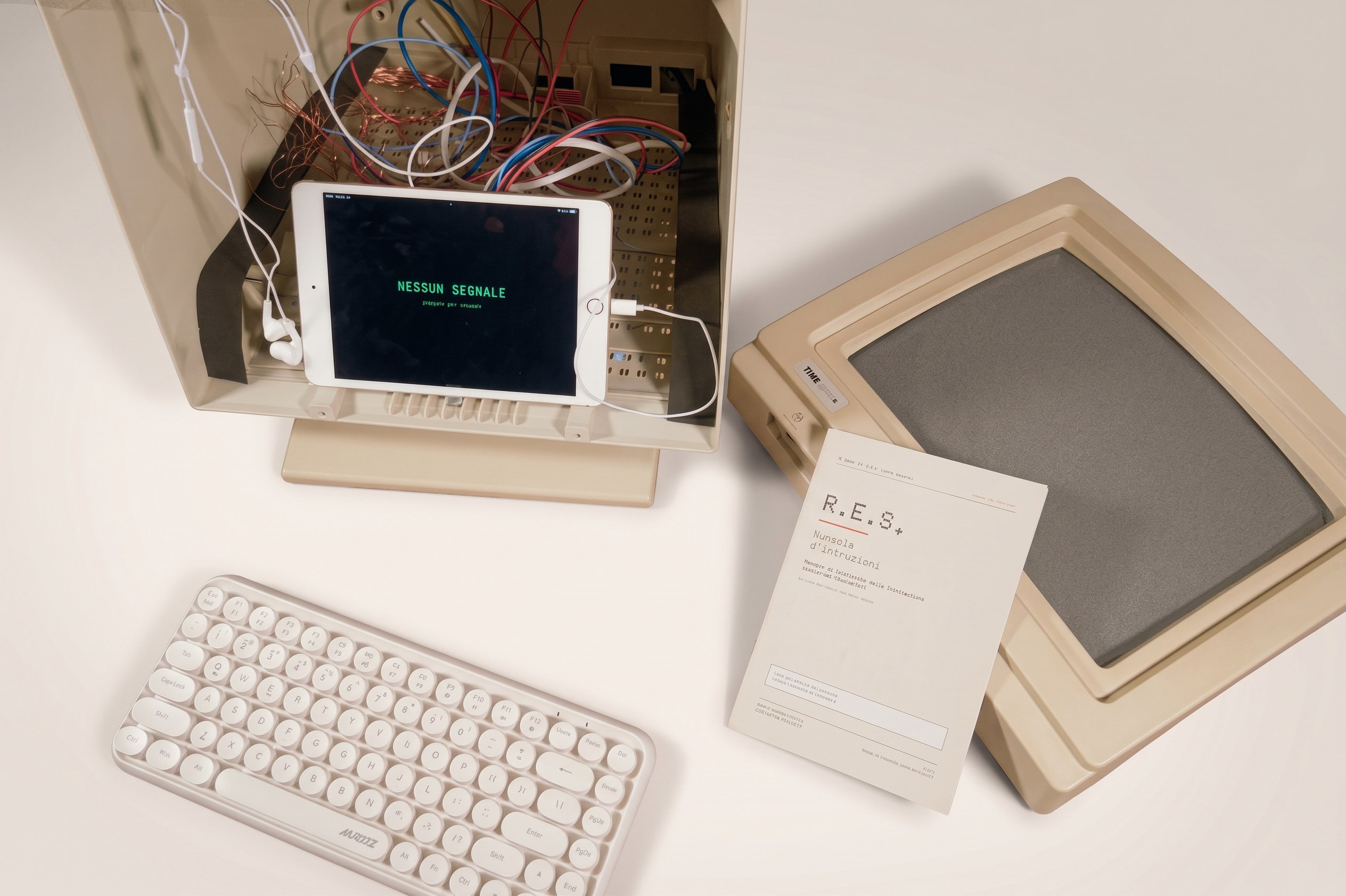 The installation from above: the monitor opened like a box, the iPad mini inside showing NESSUN SEGNALE among coloured wires, the white keyboard, the printed manual and the monitor's front frame set aside.